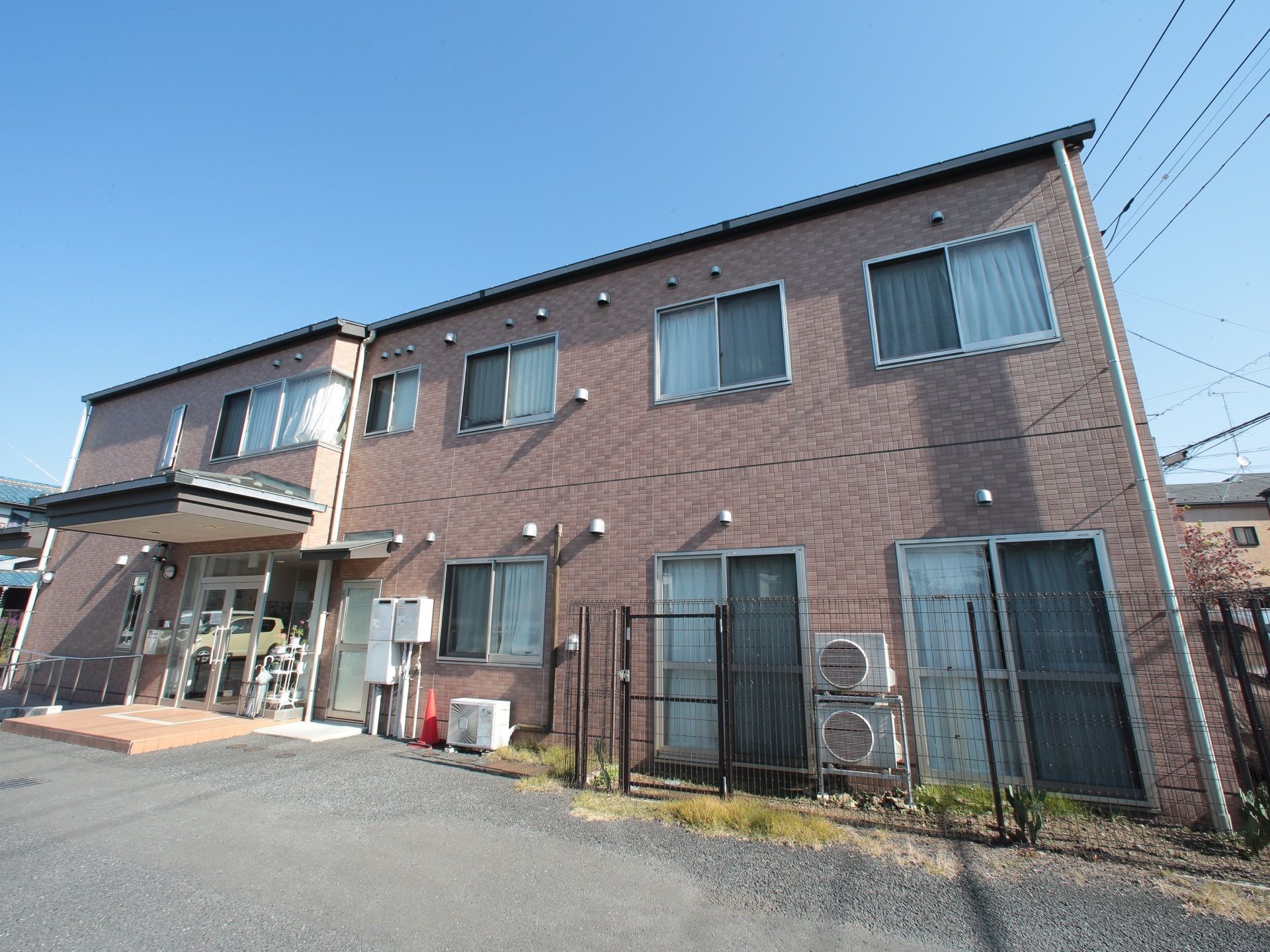 ブラウン色の2階建ての建物。玄関には庇があり、横にスロープがついており、玄関前は広い駐車場になっている。