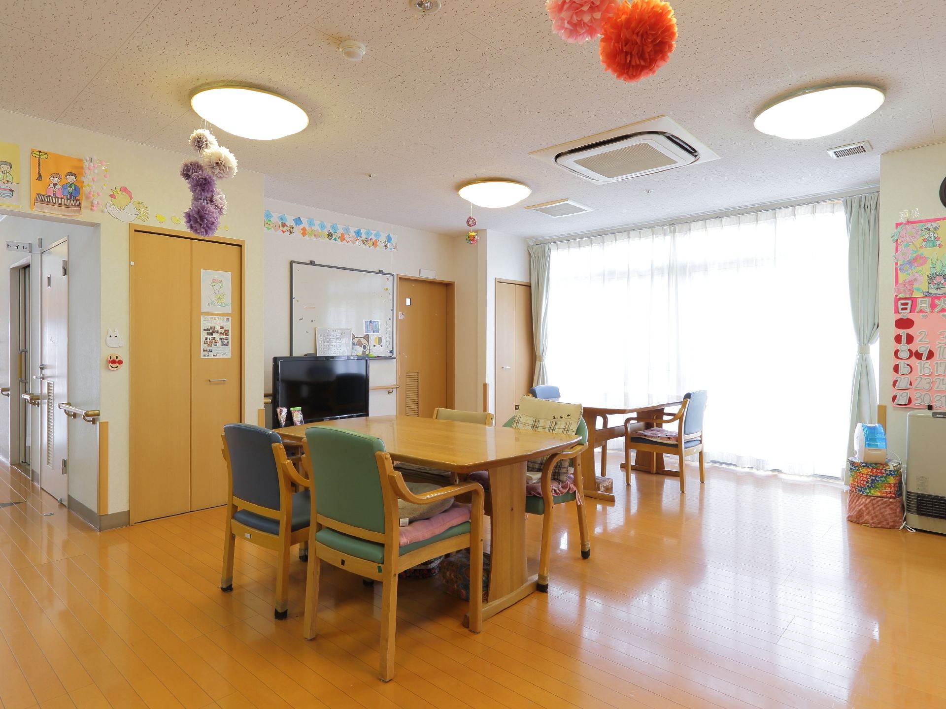 白い天井と壁に木目調の床。横には、一面のガラス戸。中央にはテーブルと椅子が並べてあり、中央の壁側にはテレビが置いてある。