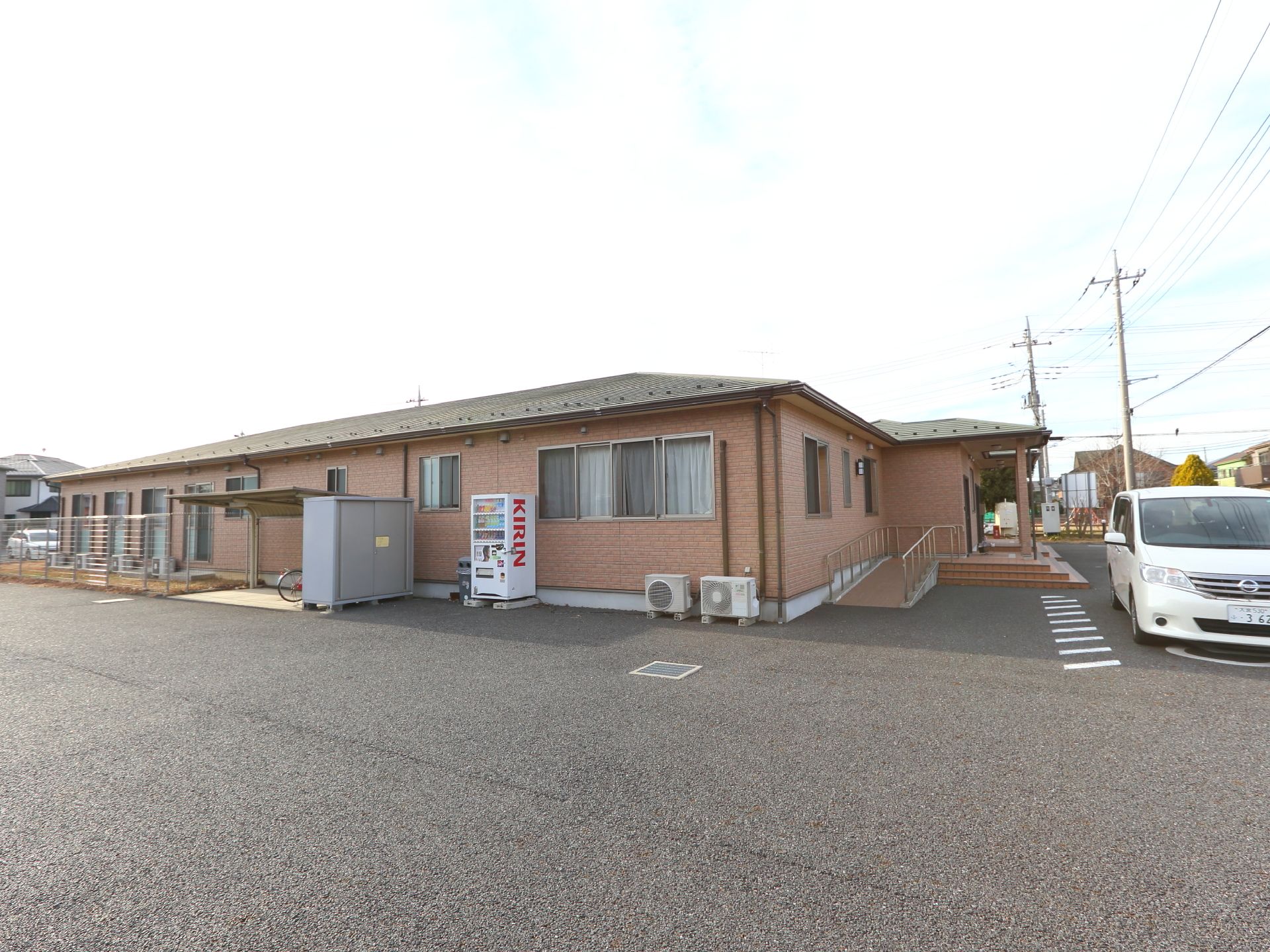 広々とした敷地内に緑の瓦屋根にレンガ調の外壁の木造平屋建ての建物。建物の壁際には自動販売機が設置されている。