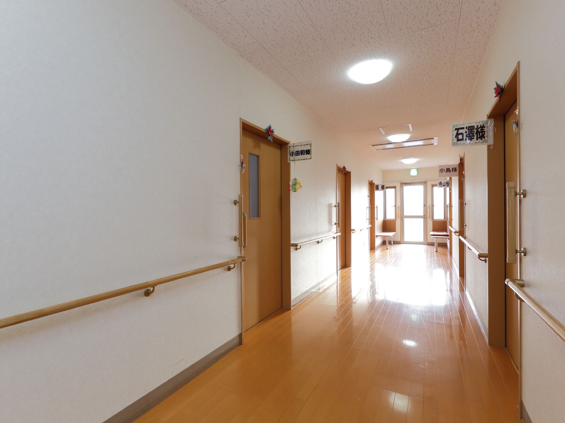 白系でまとめられた天井と壁に茶色の床仕様の通路。壁には手すりが設置され、各個室のドアの上に名前札が掲げてある。