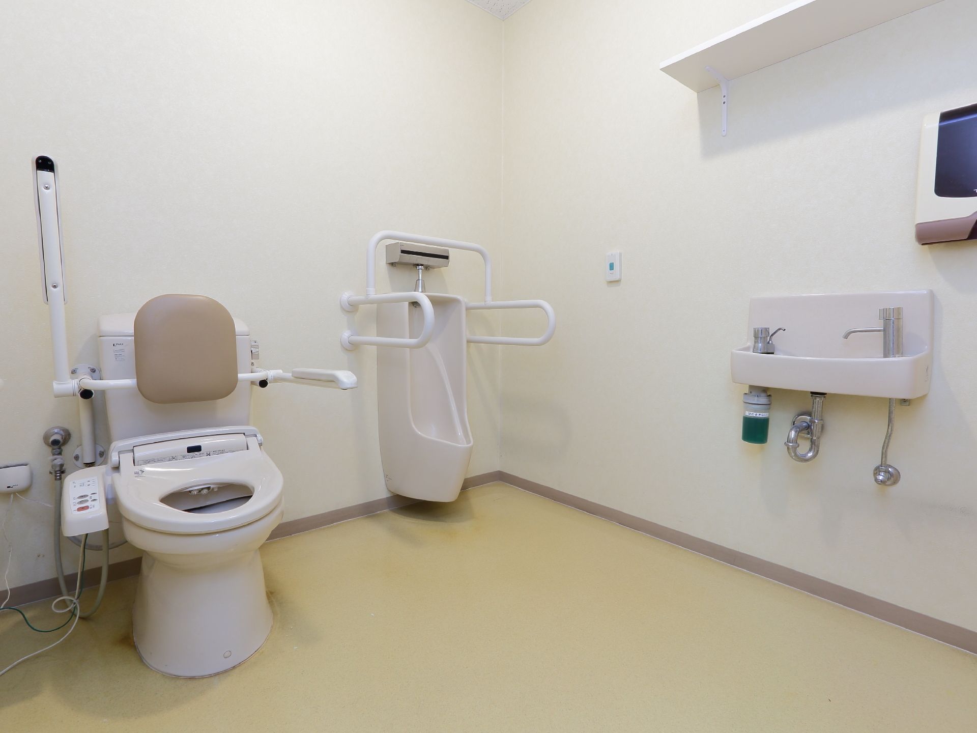 白色の壁にクリーム色の床のトイレ。男性専用の便器と2つの便器が設置。便器の傍には手すりがあり、男性専用便器の前方の壁際には洗面台がある。