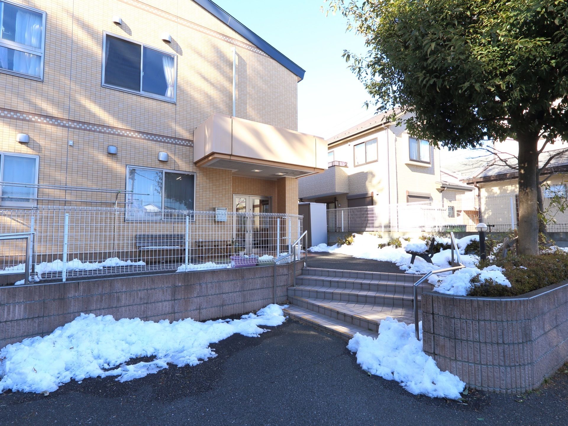 ベージュ色の２階建て建物で、周辺の住宅に馴染んでいる。手すり付きの階段を上がるとすぐ施設玄関に辿り着く。