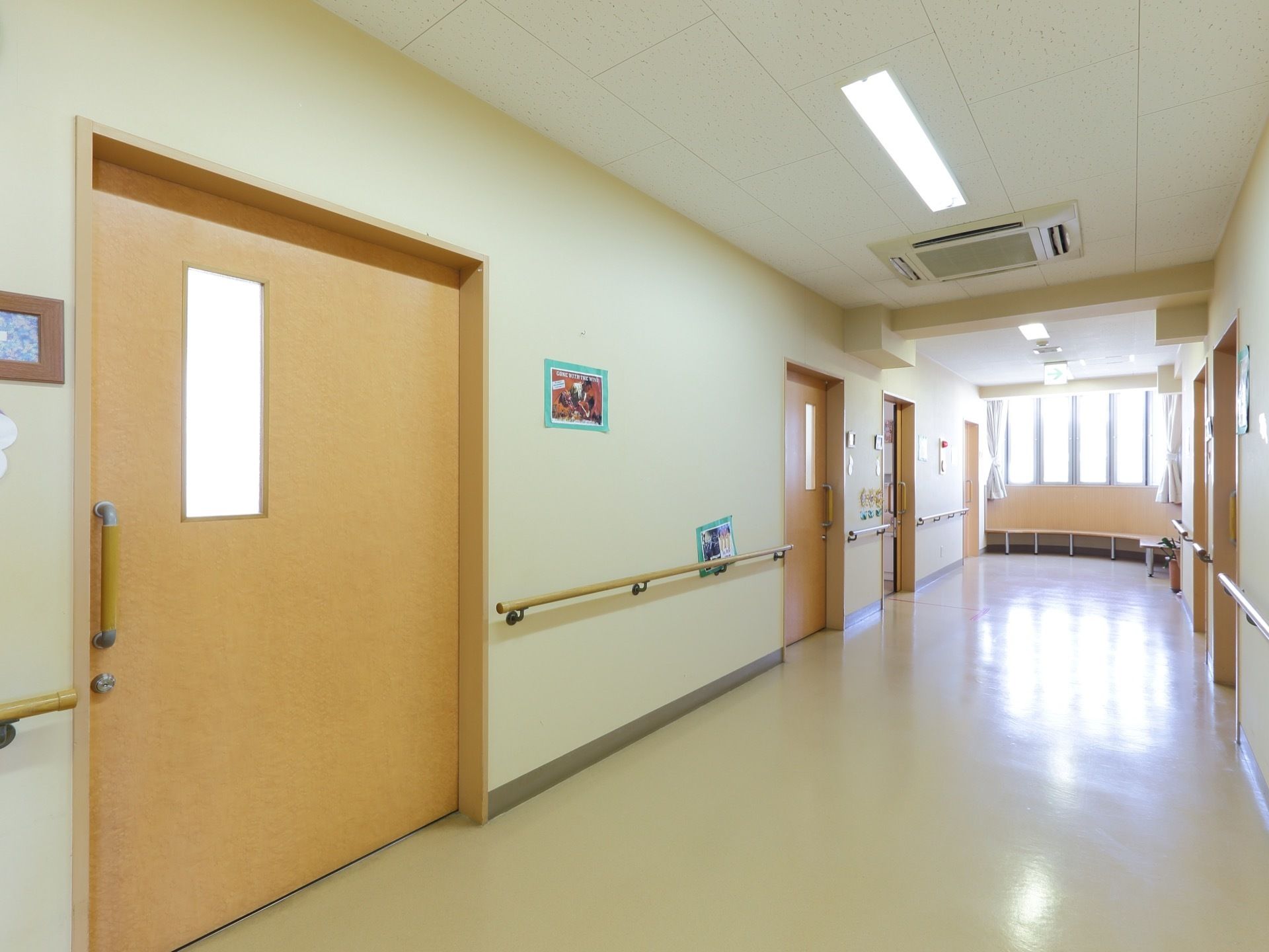 広々とした廊下には、両側に大きな引戸の取りつけられた居室が並んでいる。廊下の奥には縦長の枠のついた窓が取りつけられている。