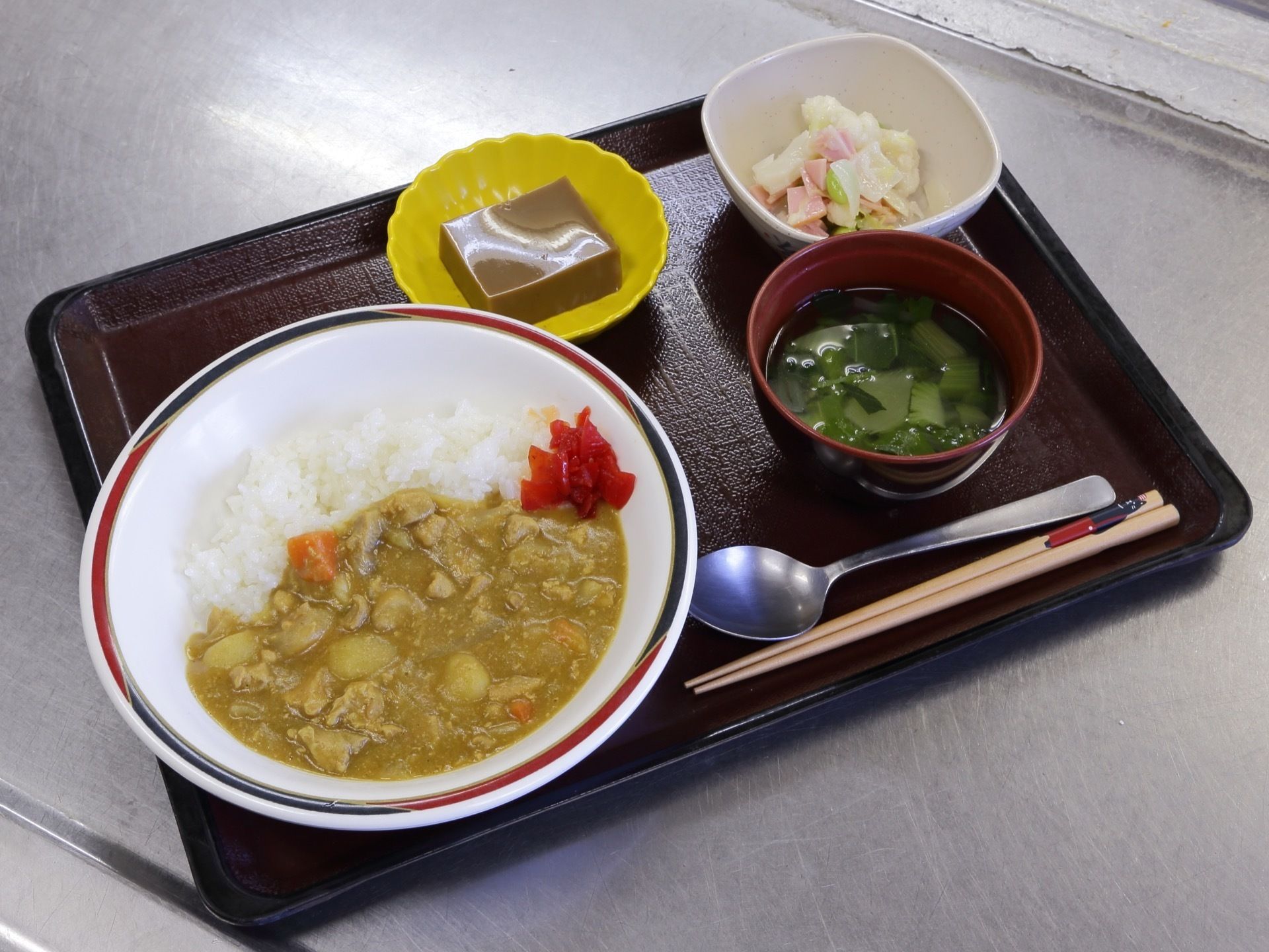 茶色いトレーの上には、カレーライスやスープなどの食事が並べられている。カレーライスには赤い福神漬けも添えられている。