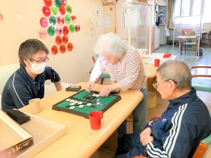 オセロを挟んで向かい合う二名の男性とオセロに手を伸ばす女性