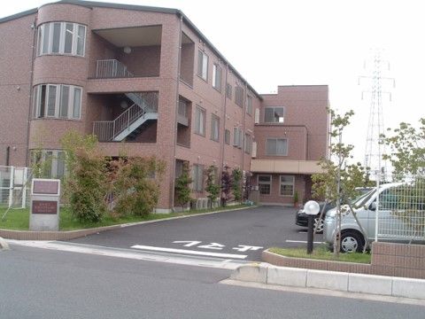 外壁がレンガ色の３階建ての施設である。外階段が設置されており、建物前に駐車スペースが設けられている。
