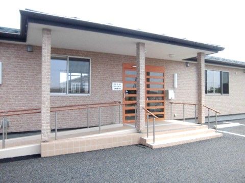 １階建ての建物で、壁の色は薄い茶色系のレンガ調だ。玄関にはひさしがあり、ドアは木とガラスの横格子で、玄関前にはスロープや階段がある。