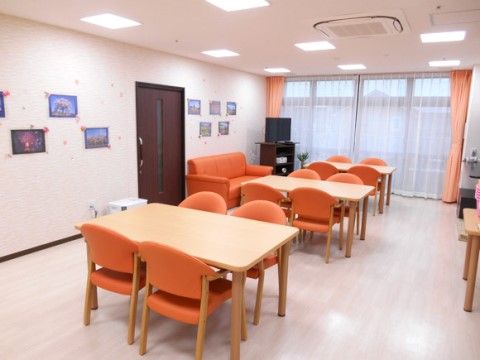 ダイニングには４人用テーブルが４卓とソファーやテレビがある。壁には絵が飾られ、カーテンや椅子、ソファーがオレンジ色で統一されている。