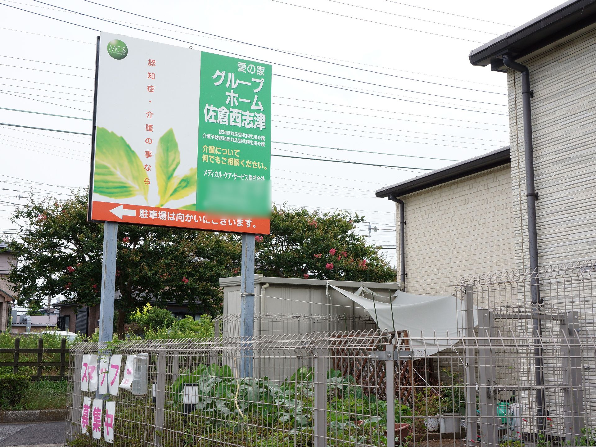 施設の駐車場への案内看板。施設の向こうには京成本線の線路が見える。周囲の環境から浮かない緑と白が基調のハデすぎない看板。