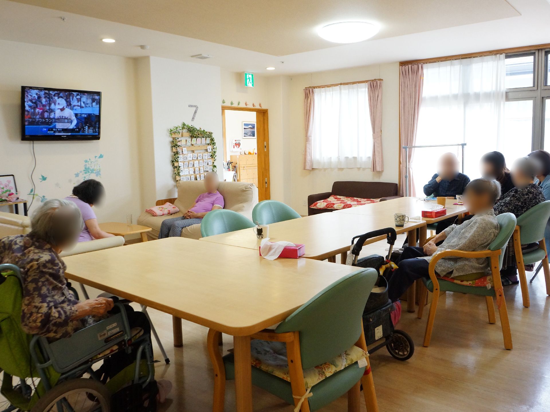 リビングルームでゆったりと過ごす利用者の様子。テレビを見たり、お茶を飲んだりされている。大きな窓からは自然の光が入ってきている。