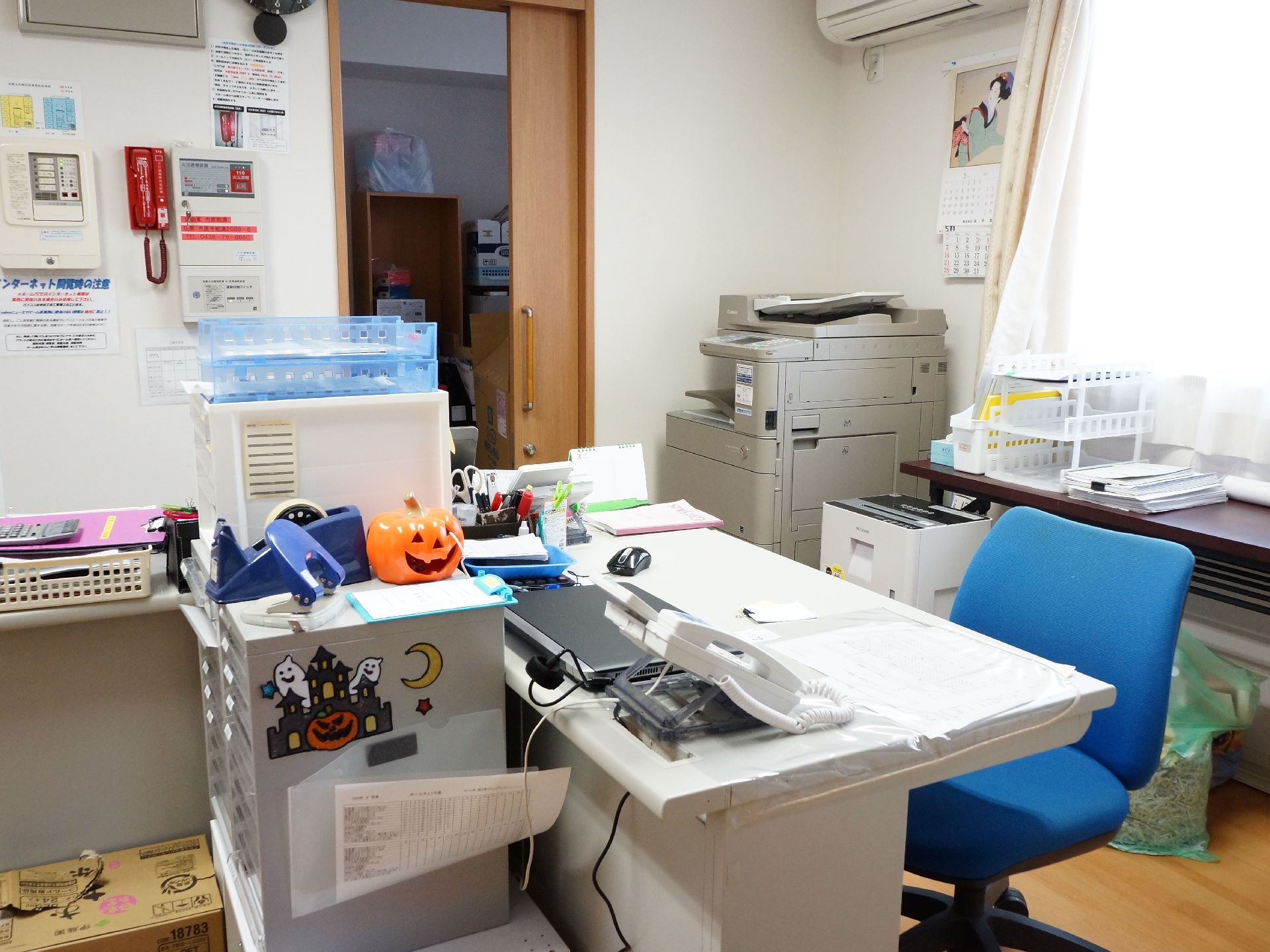 事務室に事務机一つとコピー機が設置されている。机には電話やパソコンが載っており、脇には書類ケースもある。奥にはさらに部屋がある。