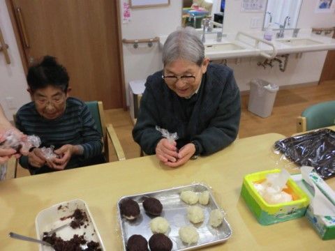 椅子に腰かけてラップでくるむようにおはぎを作る二人の男性