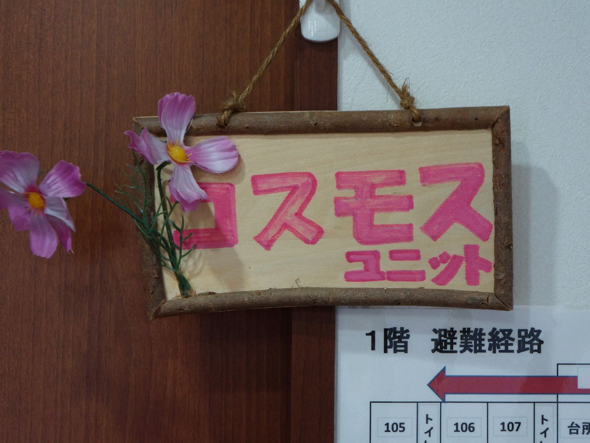 ユニット名の入った表札。手作り感のあるプレートになっており、造花のコスモスもユニット名に合わせて飾ってある。
