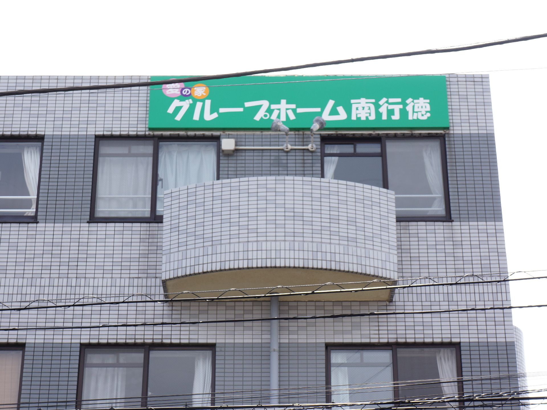外観はグレーのタイル張りで、建物最上階の正面右上に、施設の緑色の看板が設置されている。外から部屋側の窓に付けられた手すりが見える。