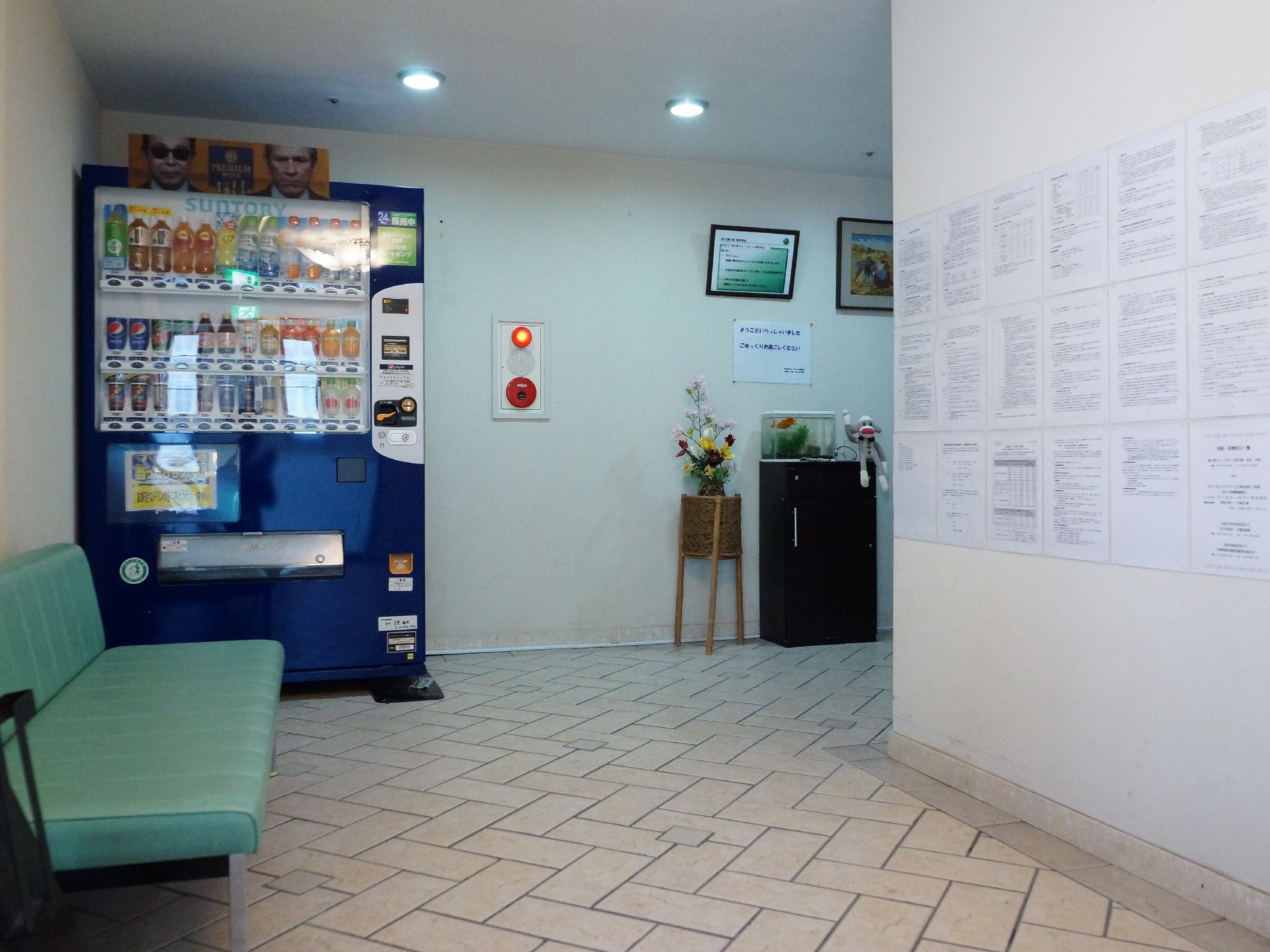 内部のL字のタイル張りの廊下の角に、飲料の自動販売機が設置されている。壁には非常ボタン、掲示物、写真、絵がある。