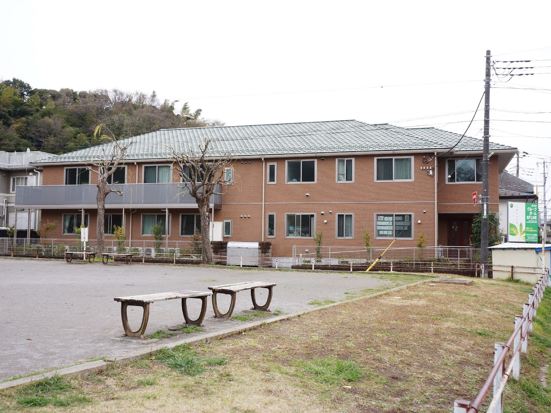 施設の道を挟んだ向かい側の公園から外観を見ると、2階部分の1部にベランダがある。敷地内の植え込みの木は背が低く、ごみ捨て場がある。