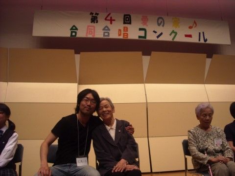 合唱コンクールの壇上で椅子に座り、肩を組んで記念写真を撮る二人