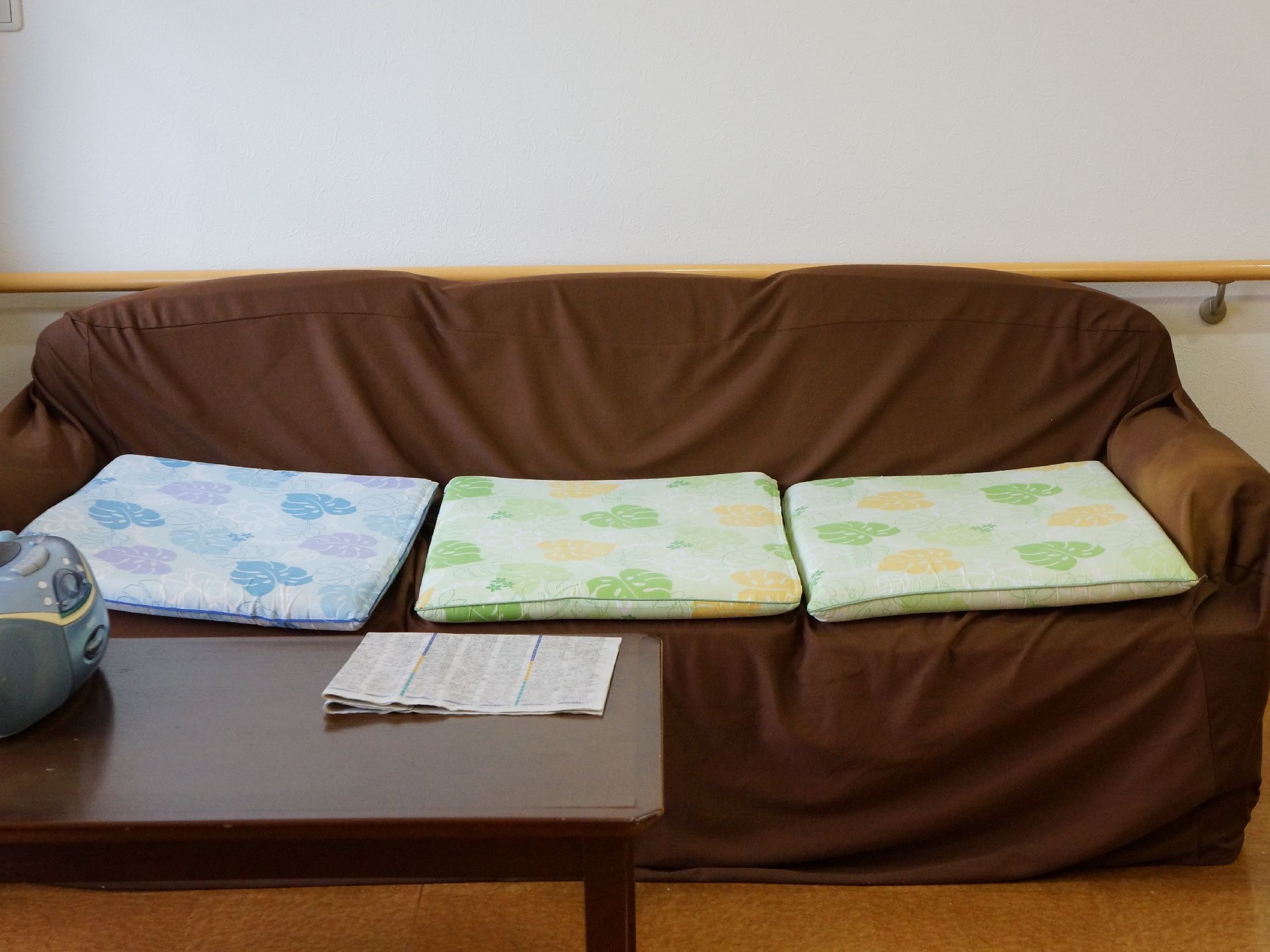 壁儀差に置かれた茶色のカバーの掛けられたソファ。3人掛けで、座布団が並べられている。テーブルの上には新聞が置いてある。