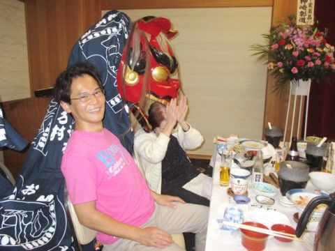 ピンクのTシャツを着た男性とその奥で獅子舞に噛みつかれる女性