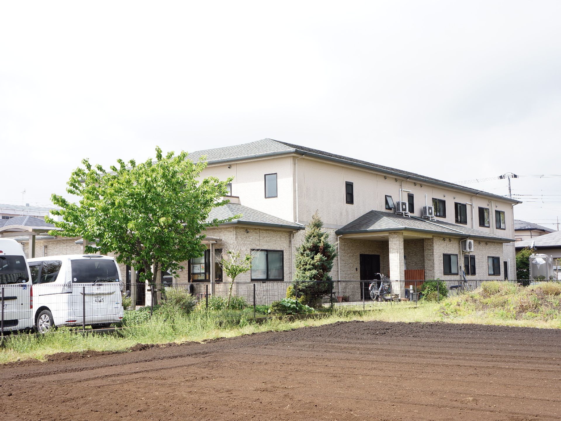 施設の建物は2階建てで、周りにあまり高い建物のない住宅街の立地にある。手前には畑が広がっていて、その横には車が停められている。