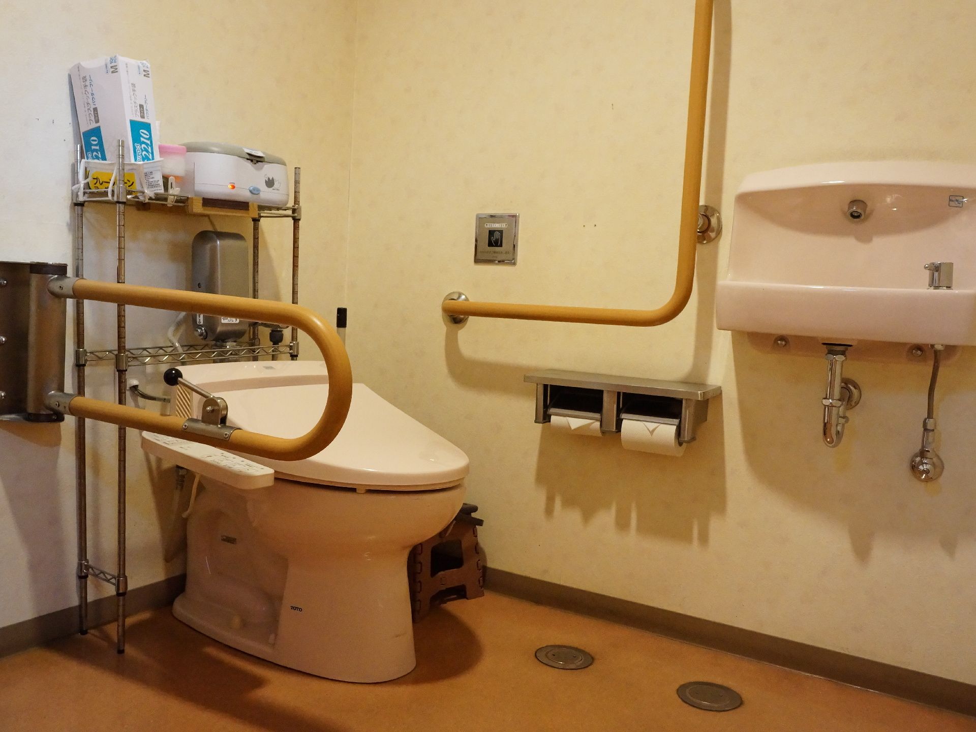洋式トイレの壁に手すりが設けられており、背後に衛生用品が置いてあるスチールラックがある。トイレットペーパーは2つセットされている。
