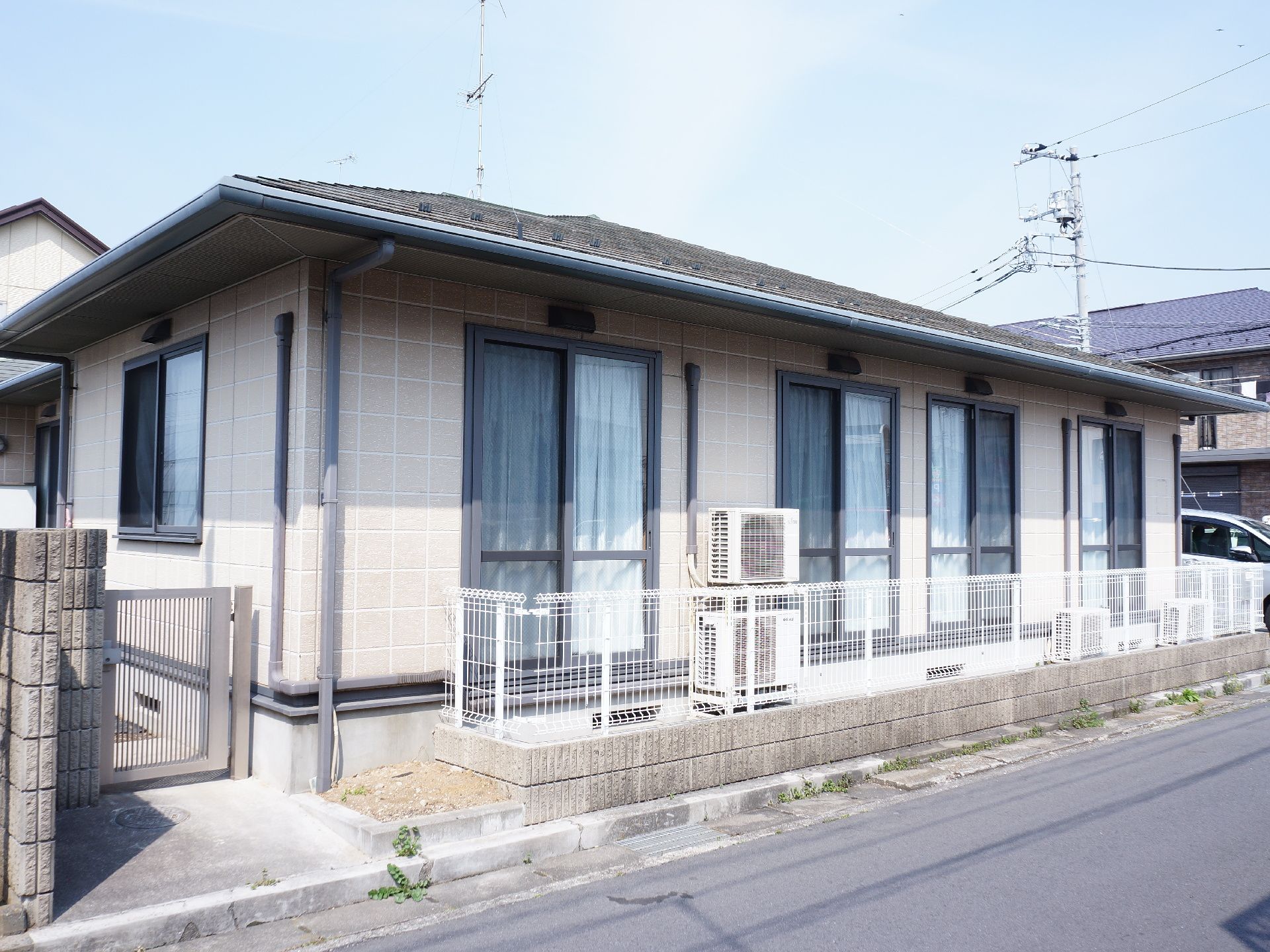 住宅街の狭い通り沿いに建っている施設の建物は1階建てで、フェンスで囲われており、脇に勝手口がある。淡いベージュ色で、窓が4つ並んでいる。