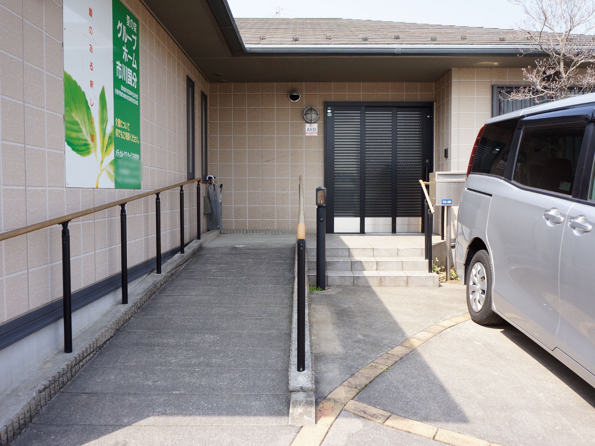 施設の玄関に向かって階段3段と壁沿いにスロープが設置されている。手前に駐車スペースがあり、ポストや照明がある。