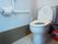 洋式トイレの壁にL字に手すりが設置してあり安全に使用できる。木目調の腰板が貼ってある。ゴミ箱と新聞が置かれている。