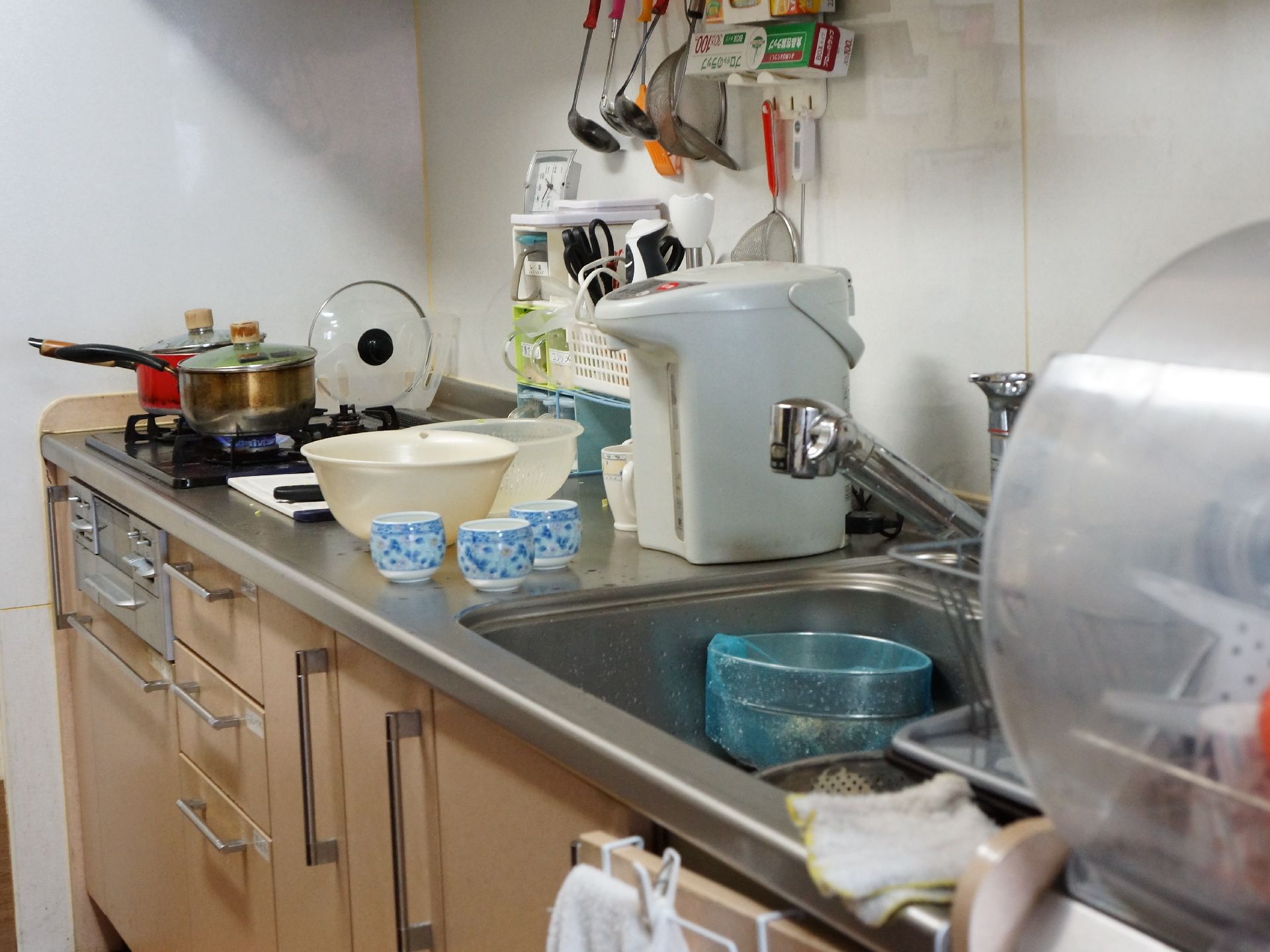 システムキッチンで調理が行われている。上には食器乾燥機、電気ポット、湯飲みや調理器具が置かれ、鍋が火にかかっている。