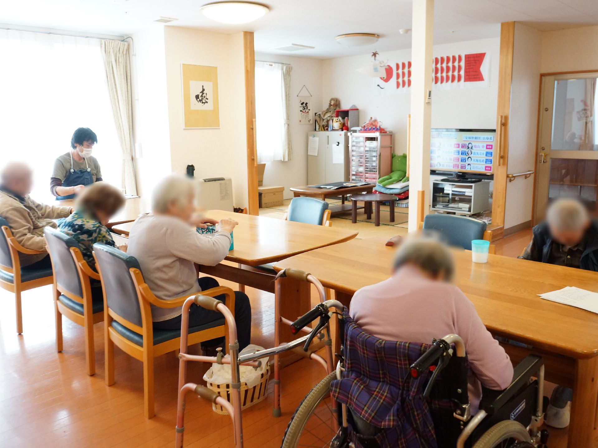 テーブルセットが3台ある広い多目的ルームは、6畳の和室が隣にありテレビがある。車椅子のお年寄りが休んでいる。