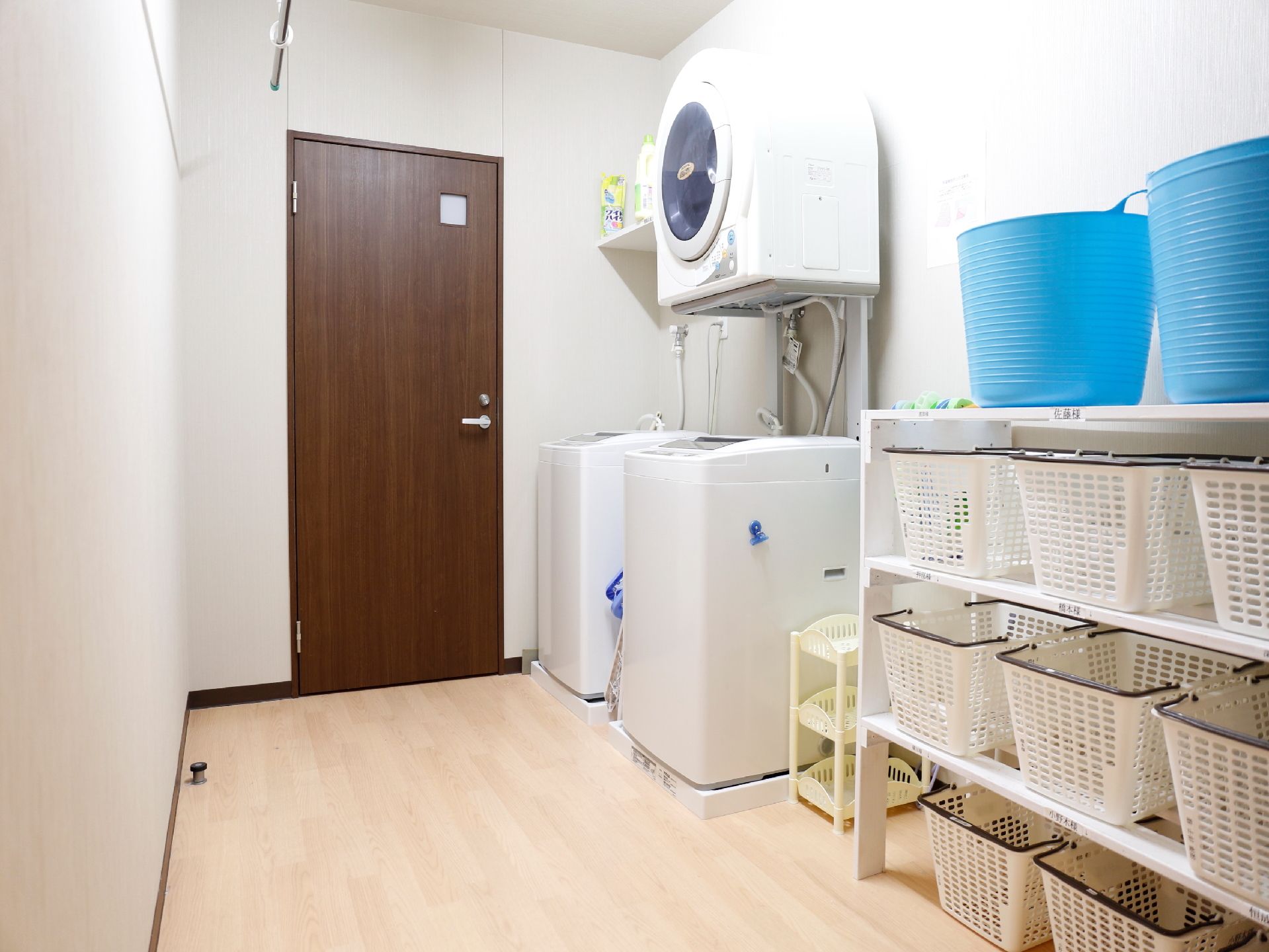 洗濯室には洗濯機が2台と、乾燥機が1台設置されている。白い棚には、入居者の名前が書かれたシールが貼ってあり、洗濯かごが置かれている。