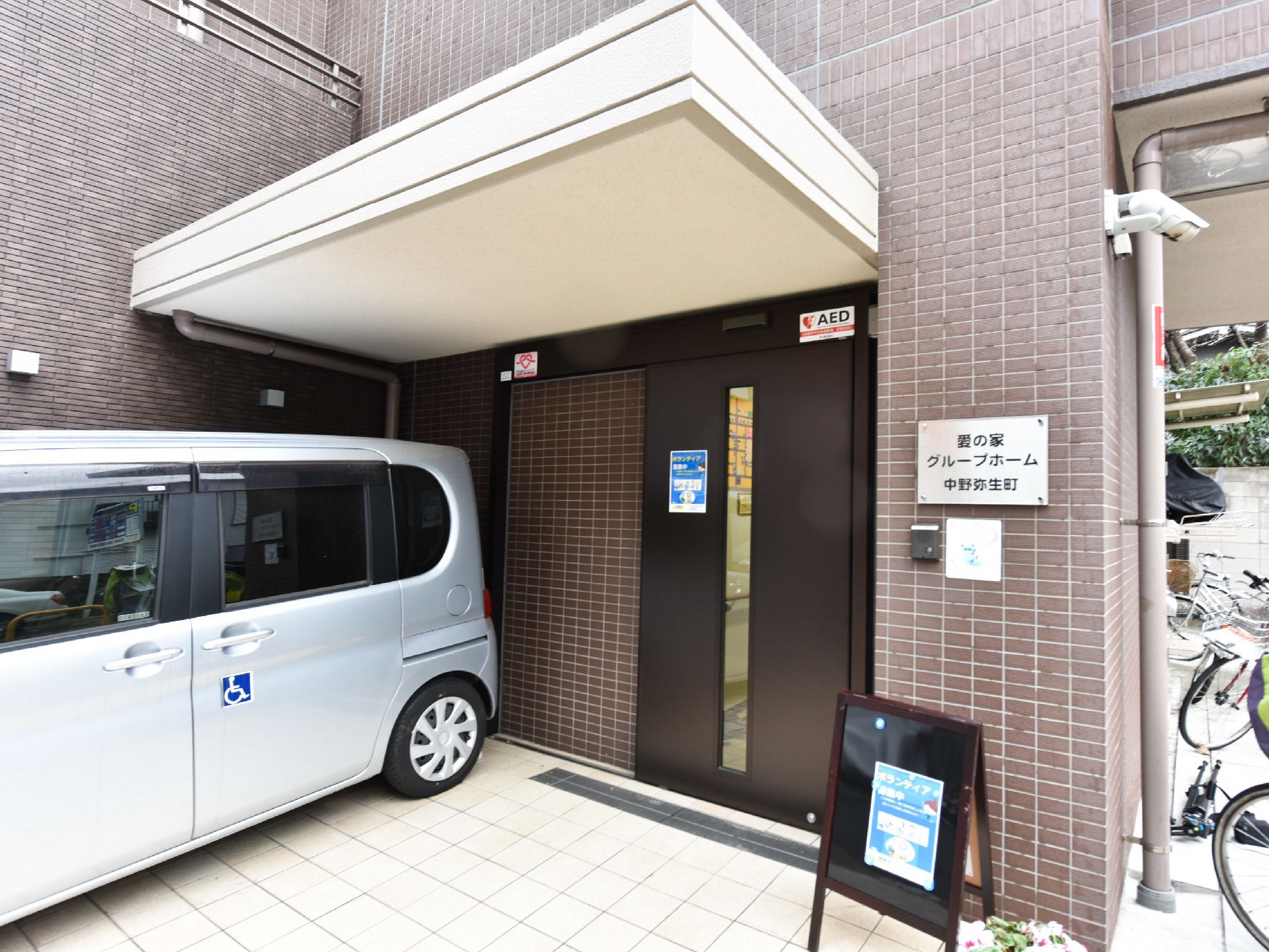 入り口の外観。ドアの隣には、車椅子を乗せることができる車が駐車している。ドアにはボランティア募集中の張り紙を貼っている。