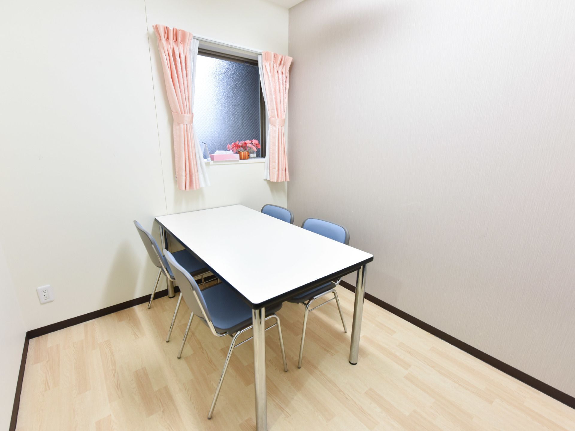 部屋には白い机と4脚の青色の椅子が置いてある。窓にはピンク色のカーテンが掛けられており、花とティッシュが置いてある。