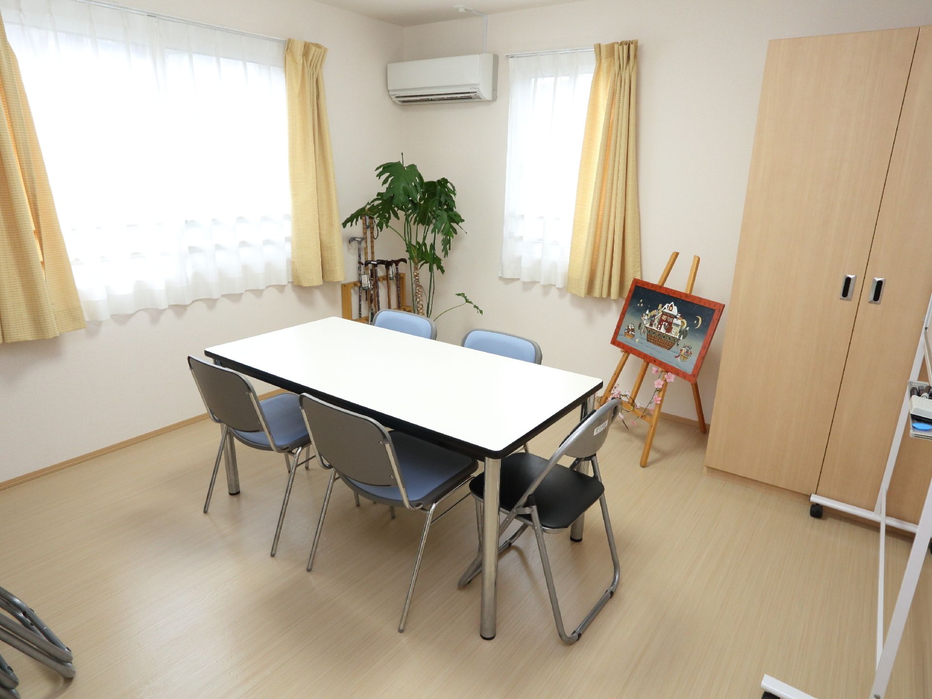 部屋の中央には、白いテーブルが置かれている。椅子は5脚ある。2か所大きな窓があり、そばには観葉植物が植えられている。
