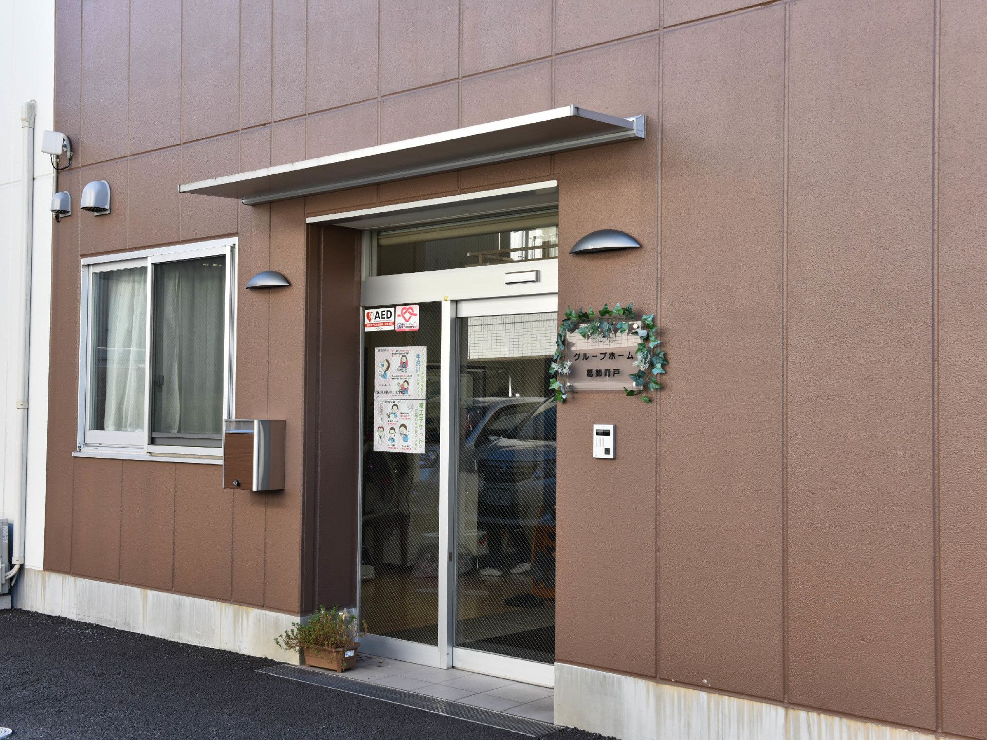施設の玄関はガラスの自動ドアで、施設名の書かれたプレートが傍に掲げられている。反対にポストが設置されている。