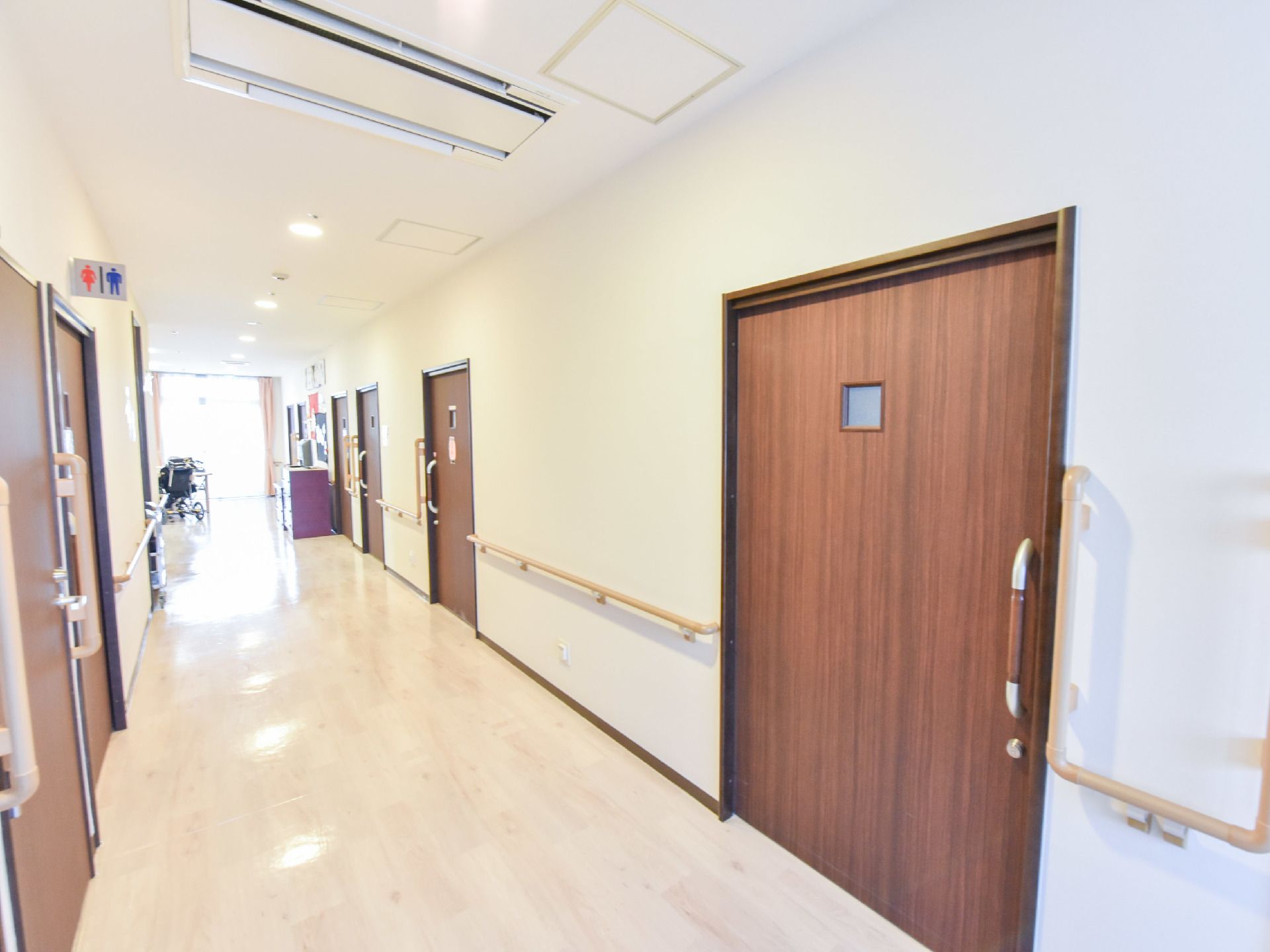 広い幅の施設内の廊下は明るく大きなスライド式ドアがいくつも並んでおり、トイレがある。予備の車椅子や手すりがある。