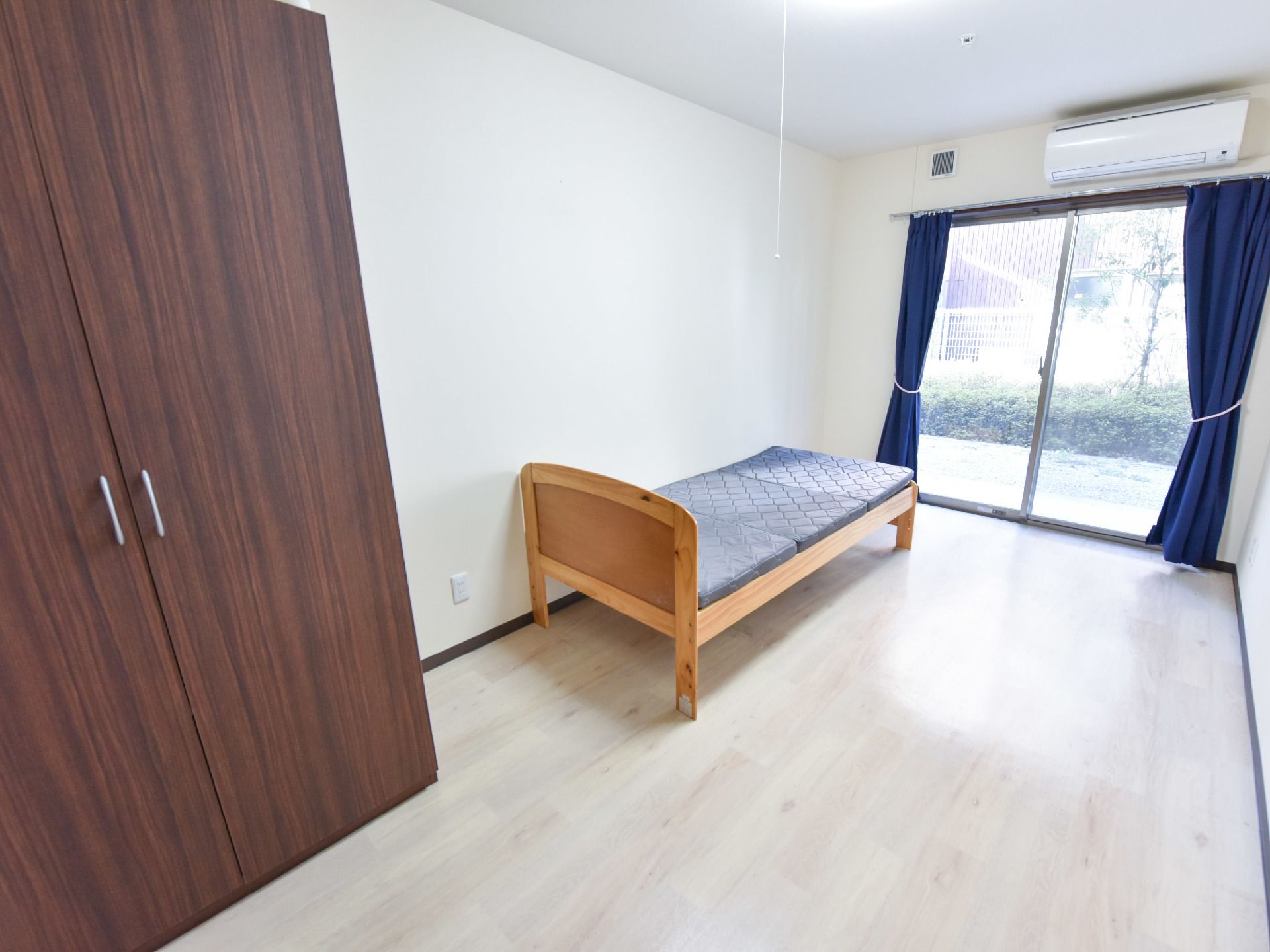 個室は一階で、シングルベッドと、据え置きクローゼット、エアコン、カーテンが付いている。外に植え込みがある。