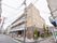商店がまばらに立ち並ぶ小さめの通り沿いに、3階建ての施設建物が建っている。2色のタイル張りで、1階部分に1台分の駐車スペースがある。