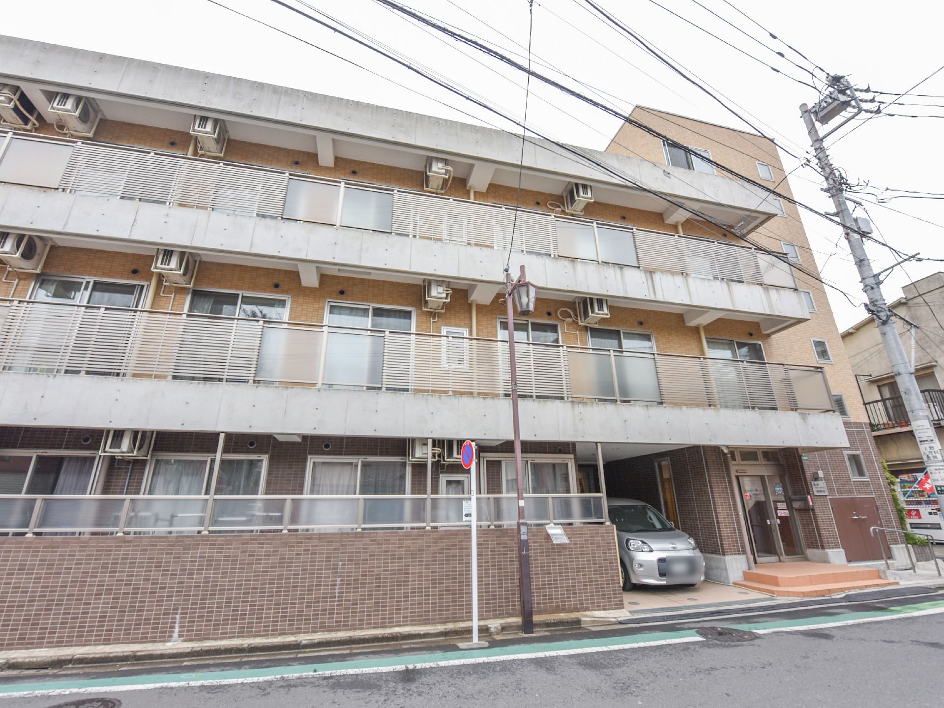 1階部分が茶色のタイル張りの建物は、歩道がある小さめの通り沿いに面して建っており、正面玄関傍に車1台分の駐車スペースがある。
