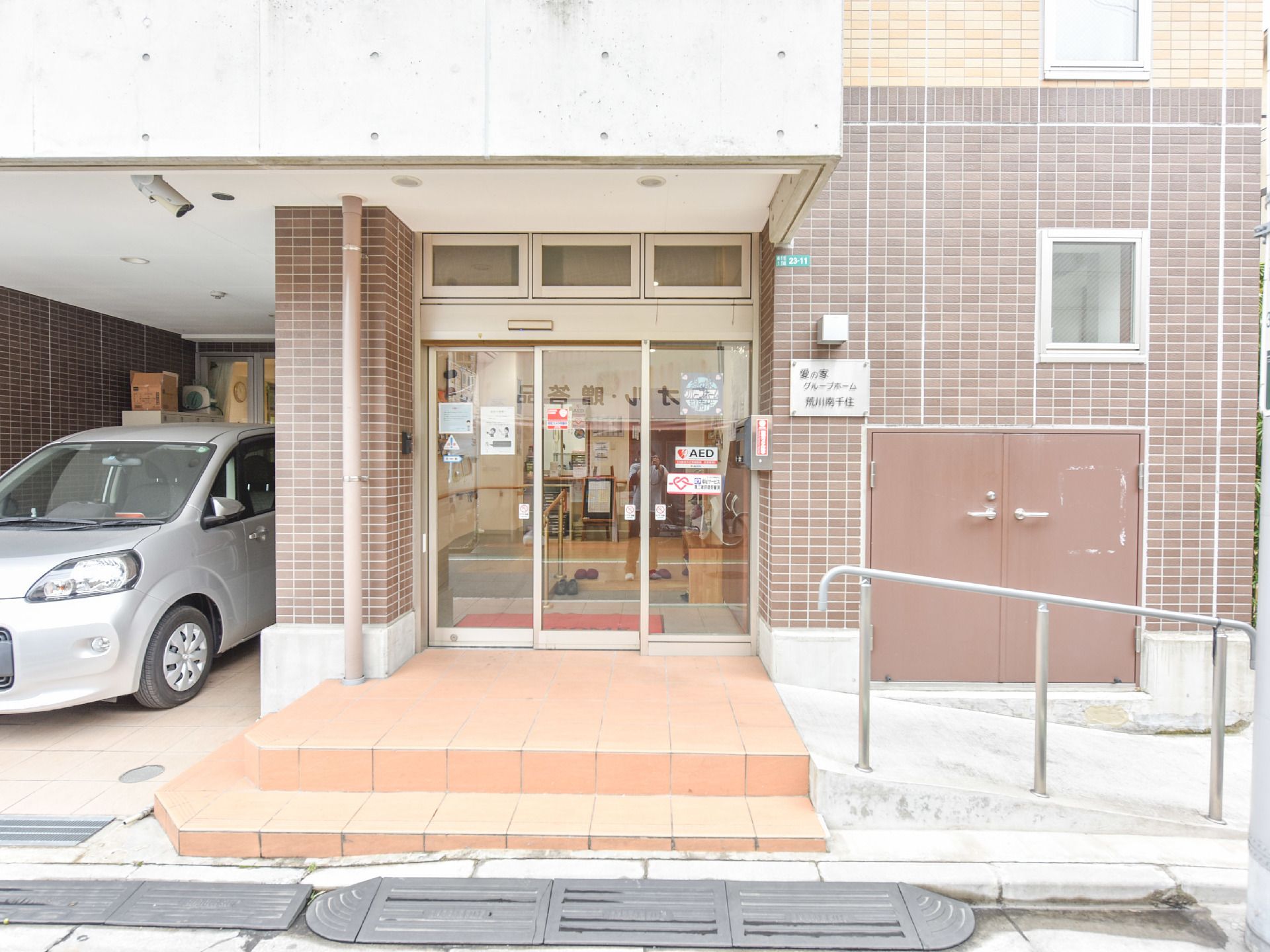 正面玄関はガラス自動ドアで、2段の階段とその傍にスロープがあり手すりが設けてある。反対側に車が1台止まっている。