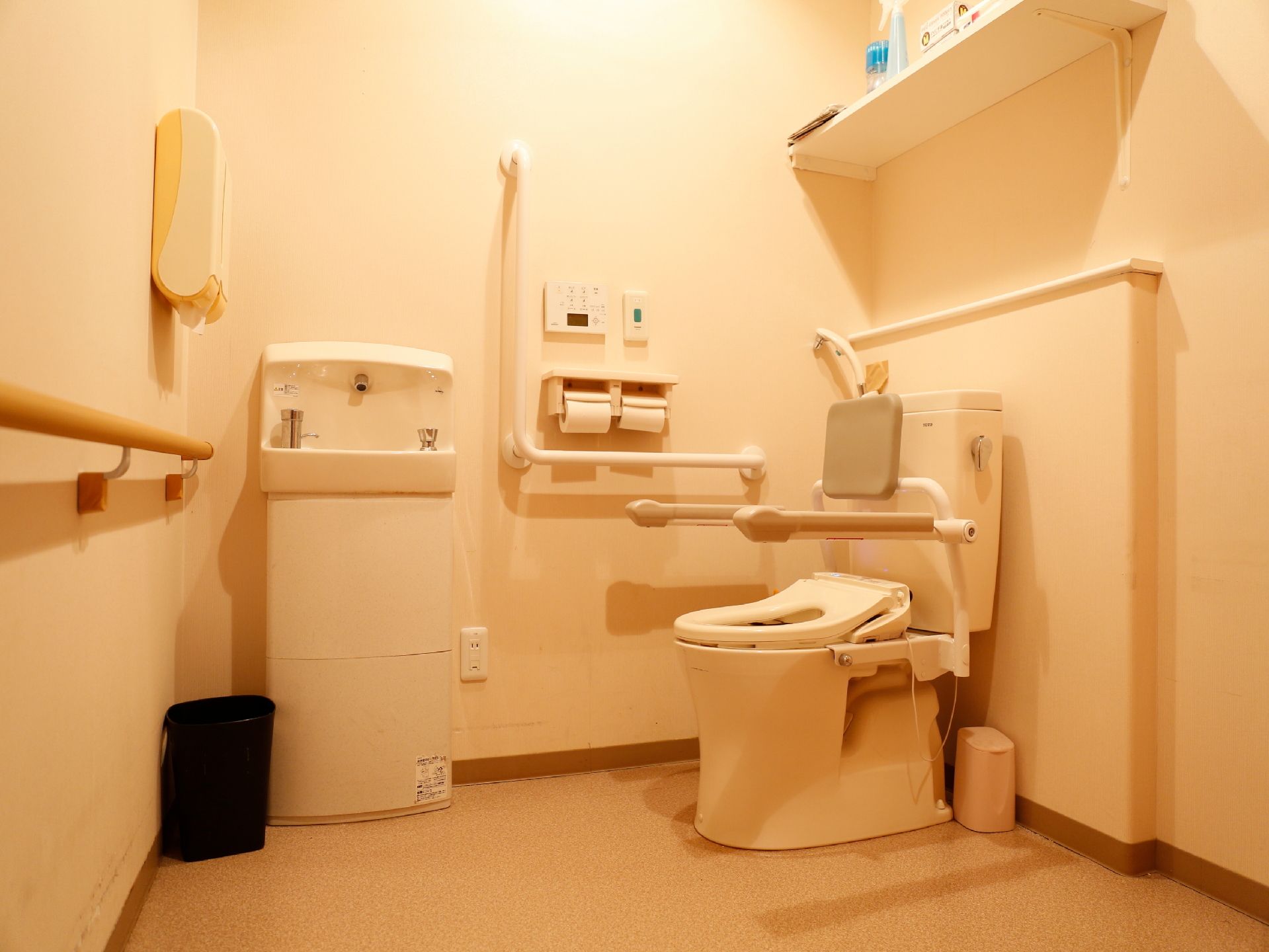 トイレスペースはとても広く、壁に洗面台、洋式便座に肘掛付き手すりと背もたれが取り付けられている。ゴミ箱がある。