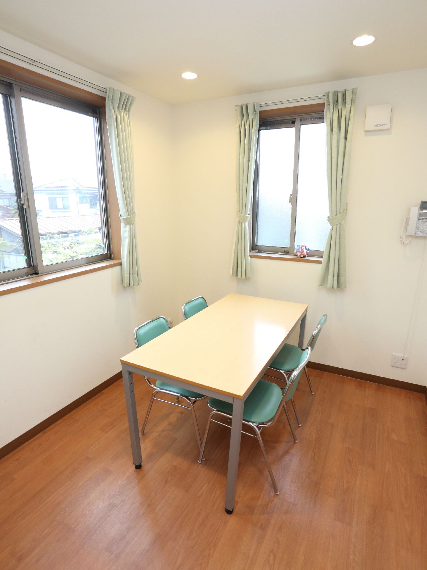 部屋の中に長机が1つと椅子が4つ置いてある。部屋には窓が2つあり、景色は開けていて開放感がある。緑色のカーテンが取り付けられている。