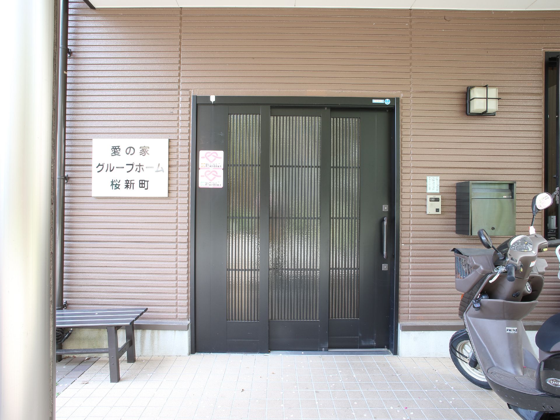 玄関ドアは3枚扉のスライド式で手動である。インターホンやポストがついている他、ベンチが置いてある。ポストの前にバイクが停められている。