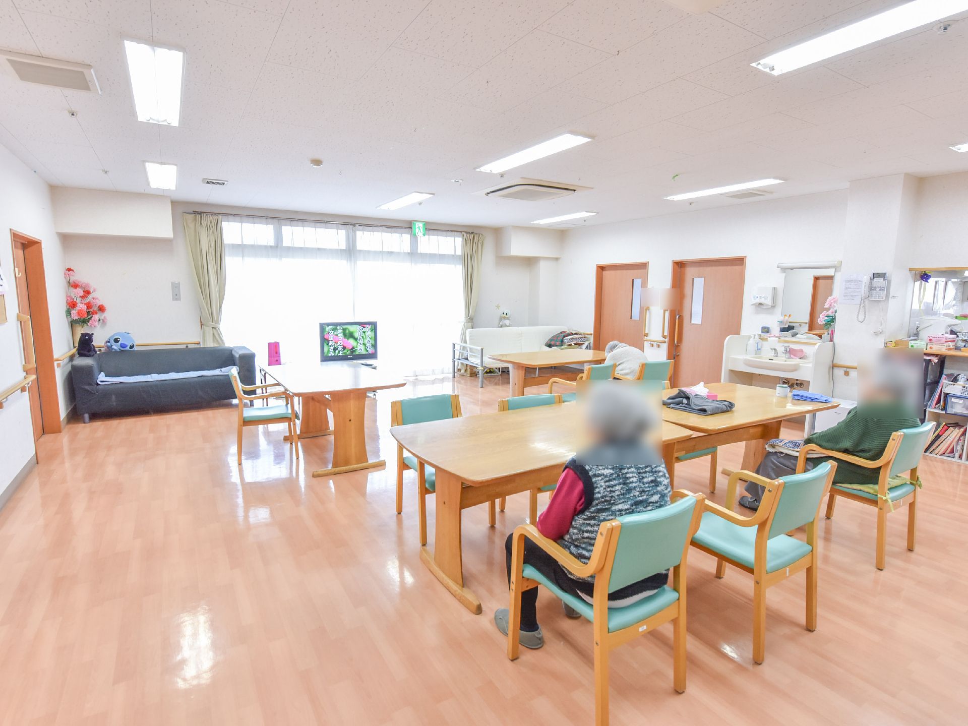 リビングの窓は大きく、上には非常口のマークがある。壁際にはテレビを挟む形で黒と白のソファーも置かれていて洗面台もあるのが見える。