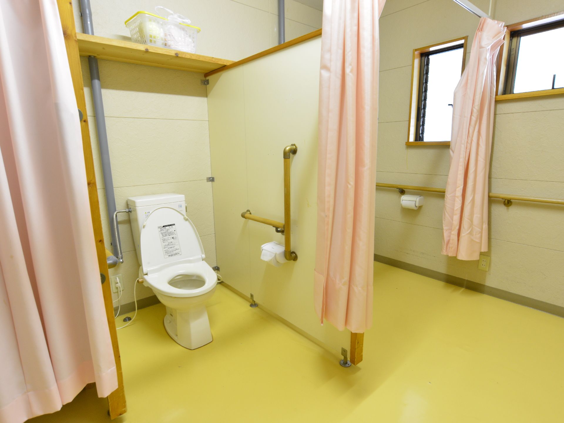 施設のトイレは白壁に黄色の床。個室に水洗トイレが設置され壁には手すりがある。個室は利用時にカーテンを閉めることができる。