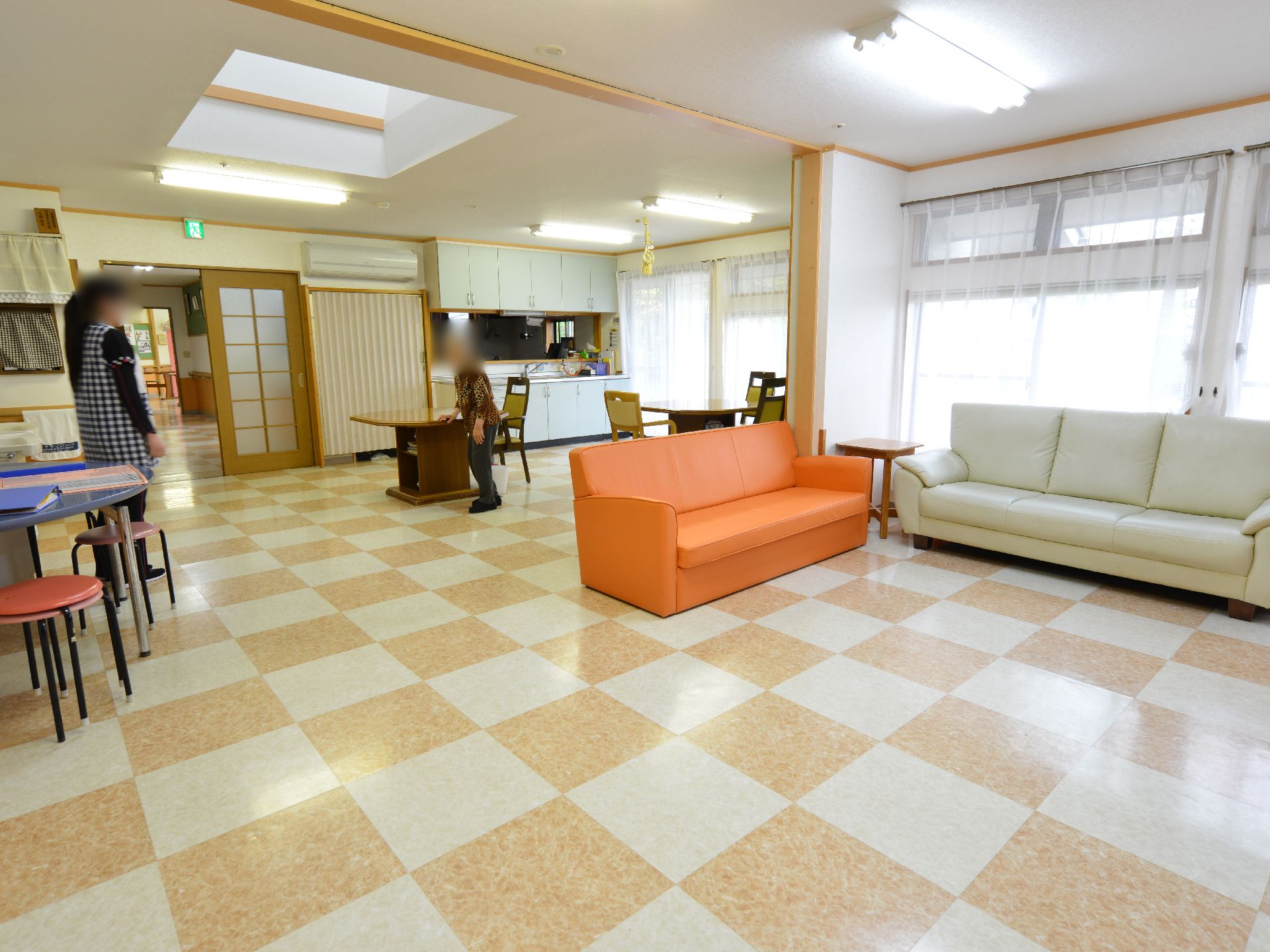 リビングは広いホールになっていてテーブルや椅子、白とオレンジのソファーなどが置かれている。窓は広く、奥にはキッチンも見える。
