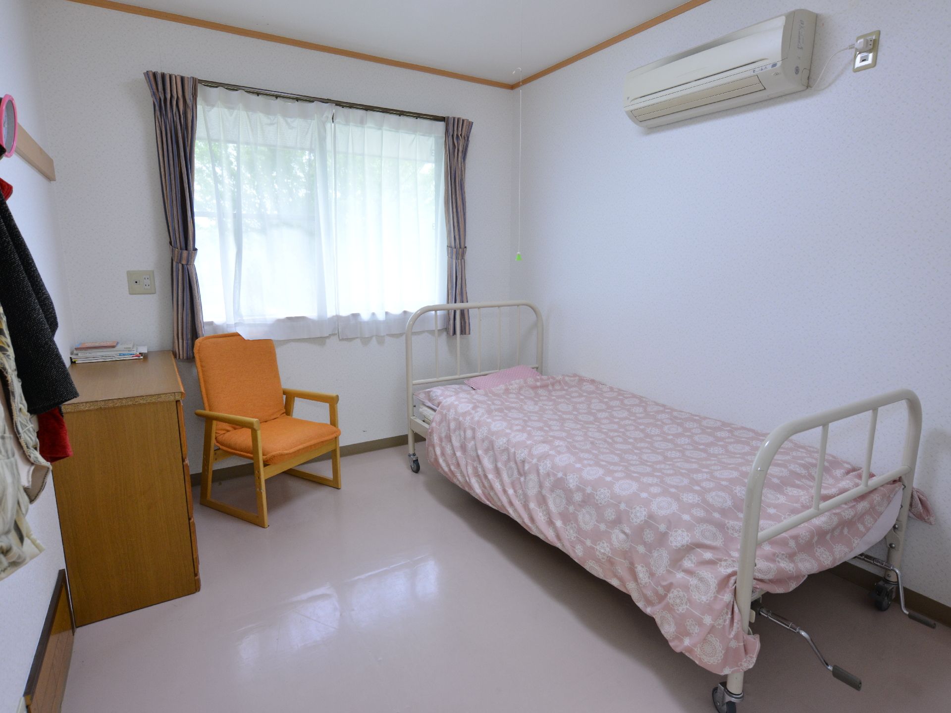 居室は白を基調とした内装。窓とカーテンがあり壁にはエアコンと服を吊るせるポールが設置されている。ベッドが1つとデスクと椅子が置かれている。
