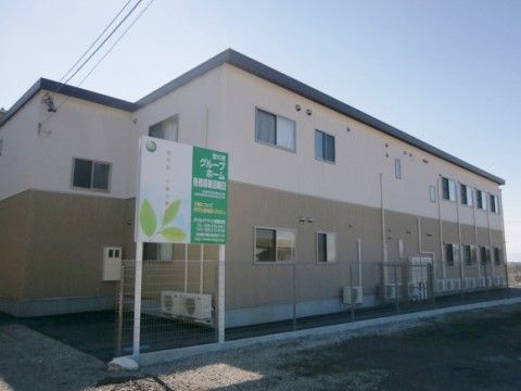下半分が濃く上半分がベージュの外壁で平らな屋根が設けられた２階建の建物は、敷地内が舗装され、境界にはフェンスがある。