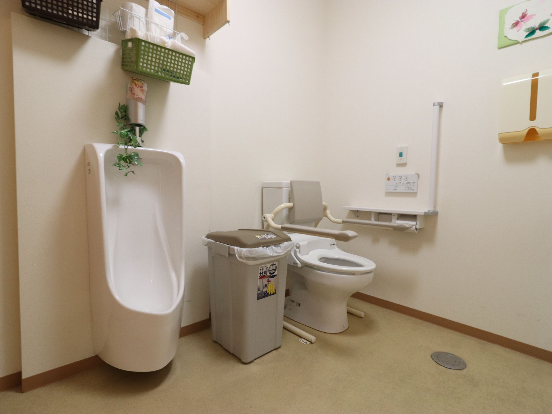 トイレは白壁にグリーンの床。男性用小便器と水洗トイレが設置されている。大便器には手すりと背もたれが外付けされている。
