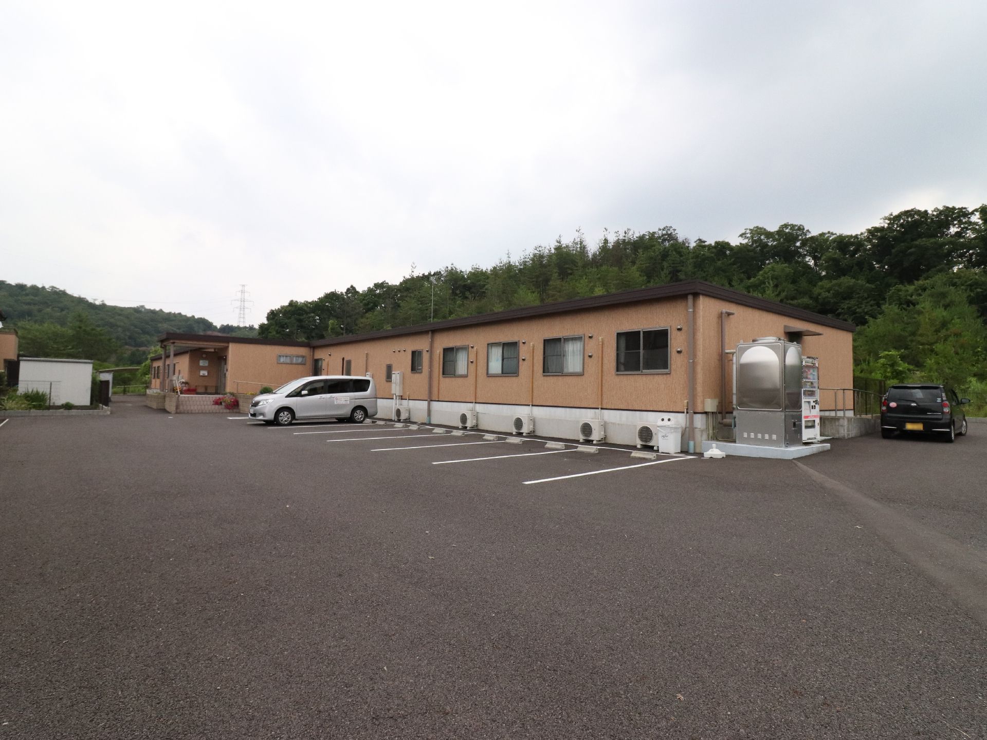 緑に囲まれた施設の周りは駐車場になっている。白い車と黒い車が一台ずつ停まっている。建物は一階建てで各部屋にエアコンが設置されている。